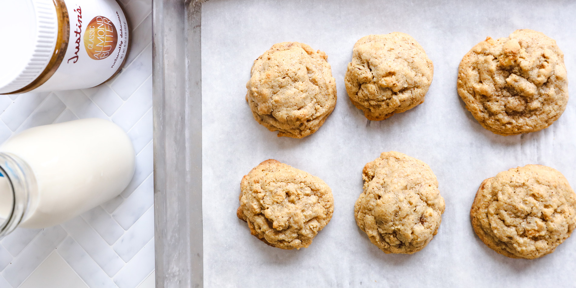 Almond Butter Granola Cookies on a parchment tray on a white background with a jug of milk beside Justin's Peanut Butter