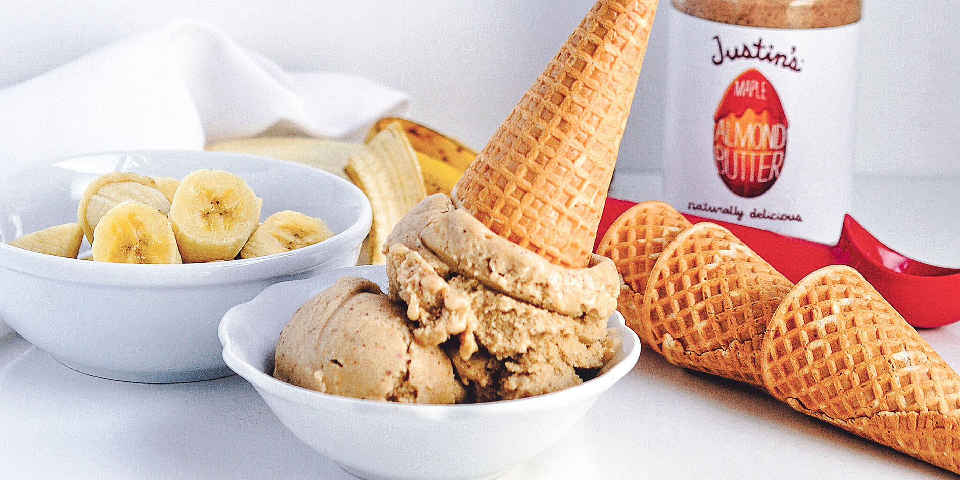 Maple Almond Butter Banana Nice Cream on a cone dunked in a small white bowl with slices bananas in another bigger white bowl