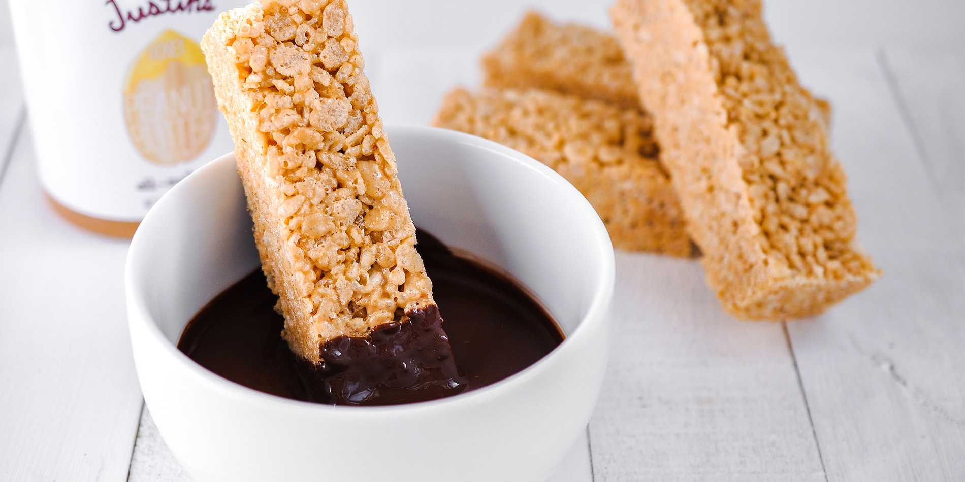 Nut Butter Rice Crispy Treats with Dark Chocolate & Sea Salt being dipped in chocolate syrup in a white bowl (front-view)