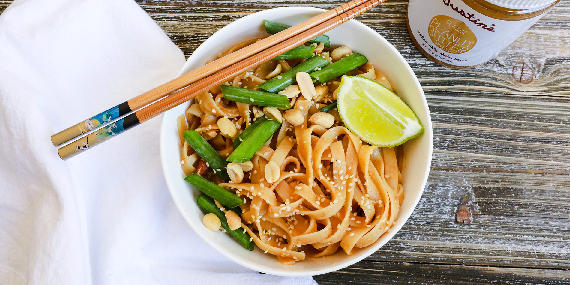 Spicy Peanut Butter Pasta with sesame seeds, peanuts, green onions and a piece of lime on a white bowl with chopsticks.