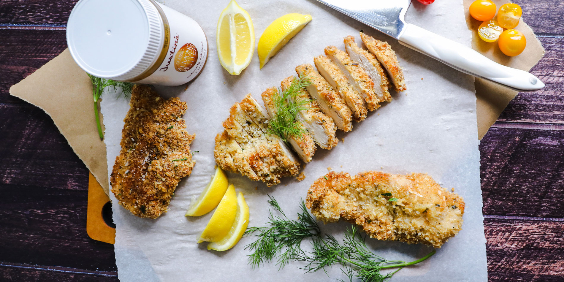 Paleo Almond Butter Crusted Chicken on parchment papers with cut baby tomatoes and lemon slices on a wooden serving tray