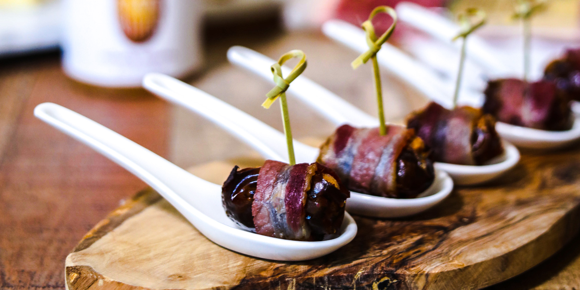 Almond Butter Stuffed Bacon Wrapped Dates on spoons on top of a wooden serving plate on a wood background