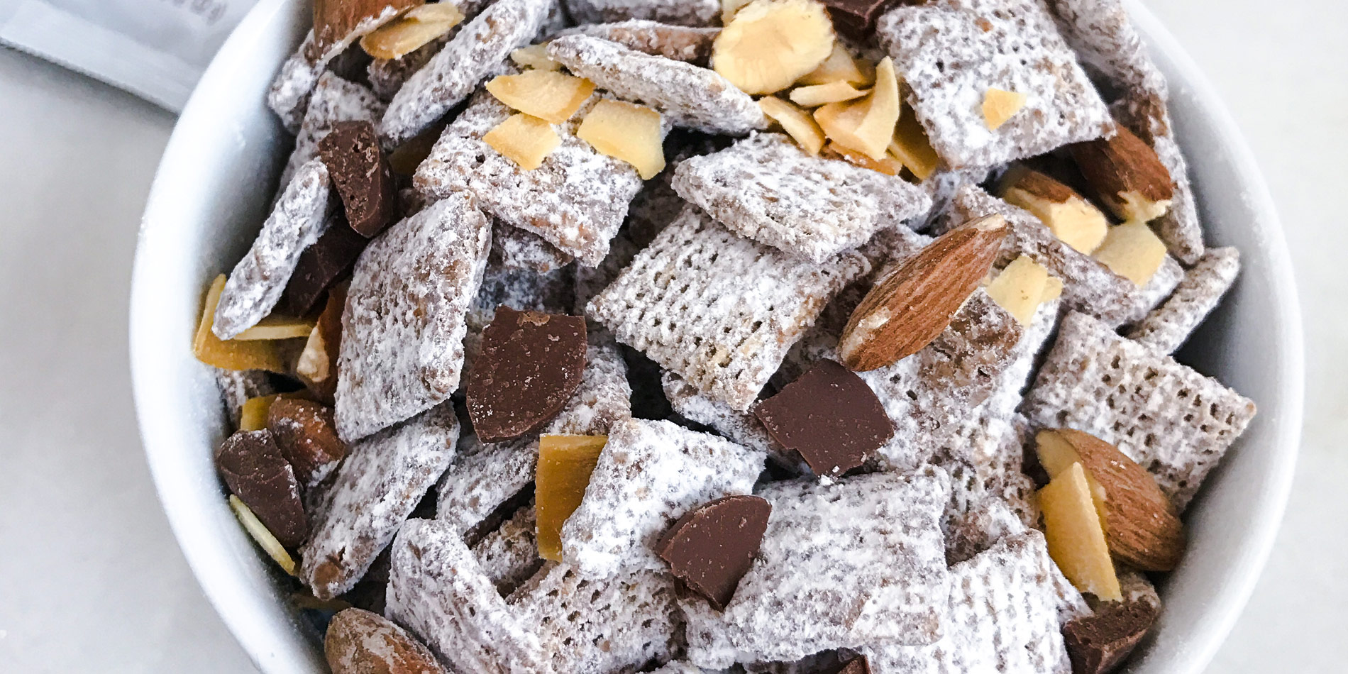 Almond Coco Nutty Buddies in a white bowl on a white background with chocolate chunks and peanut butter in measurement spoons