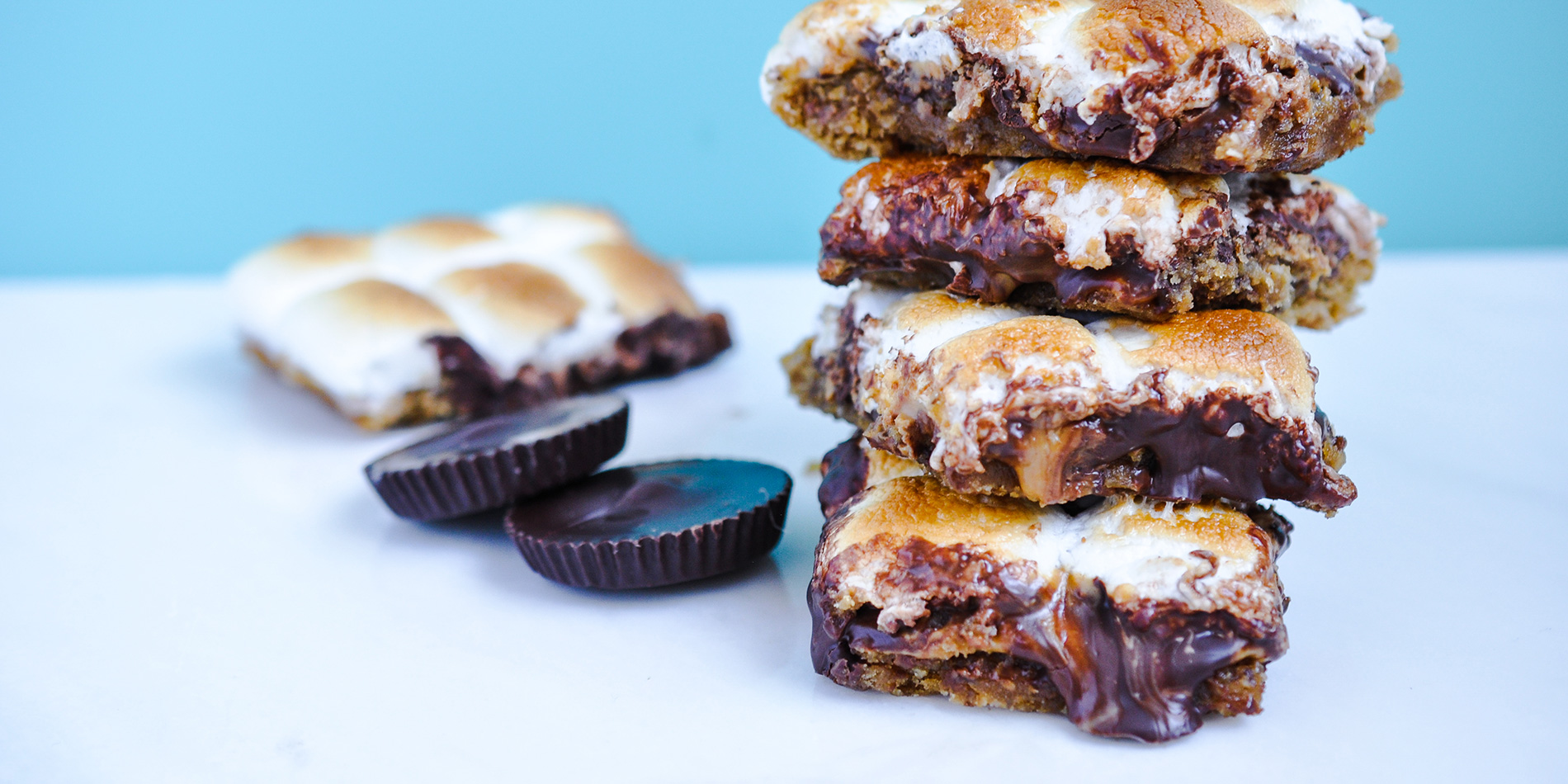 Peanut Butter Banana S'mores Bars stacked on top of each other with chocolate chips on a white top with a blue background