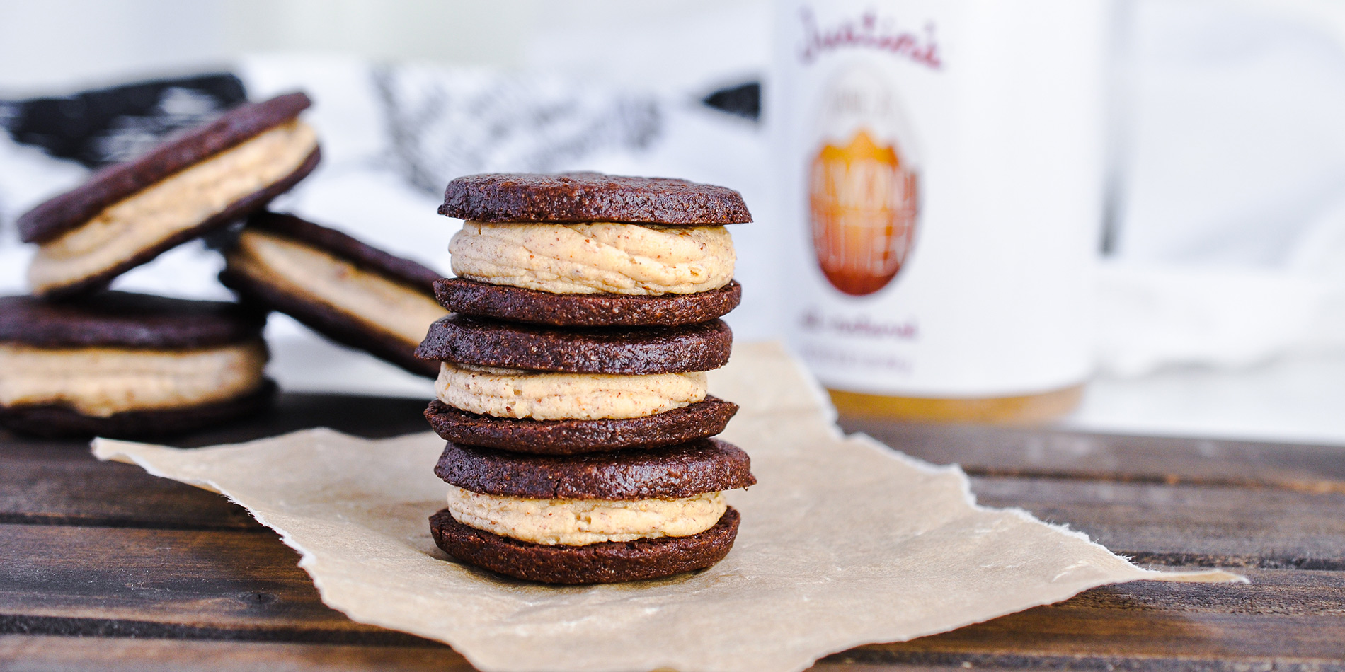 Vanilla Almond Crème Filled Sandwich Cookies stacked on parchment paper with cookies and peanut butter jar in background