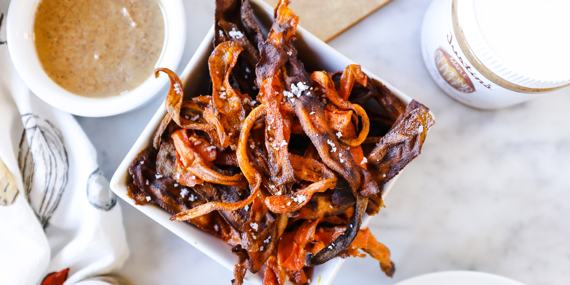 Crispy Carrot Fries with Cinnamon Maple Almond Dip with multiple sliced carrots and peanut butter jar on a white background
