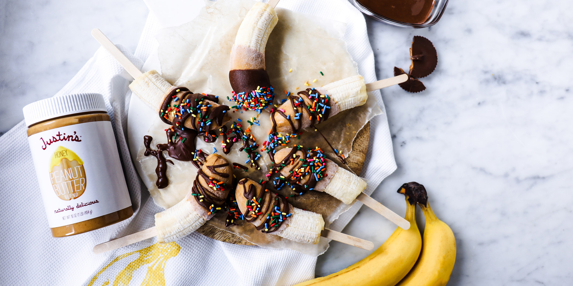 Frozen Chocolate PB Banana Pops with chocolate drizzle, rainbow sprinkles, Justin's honey peanut butter jar and two bananas