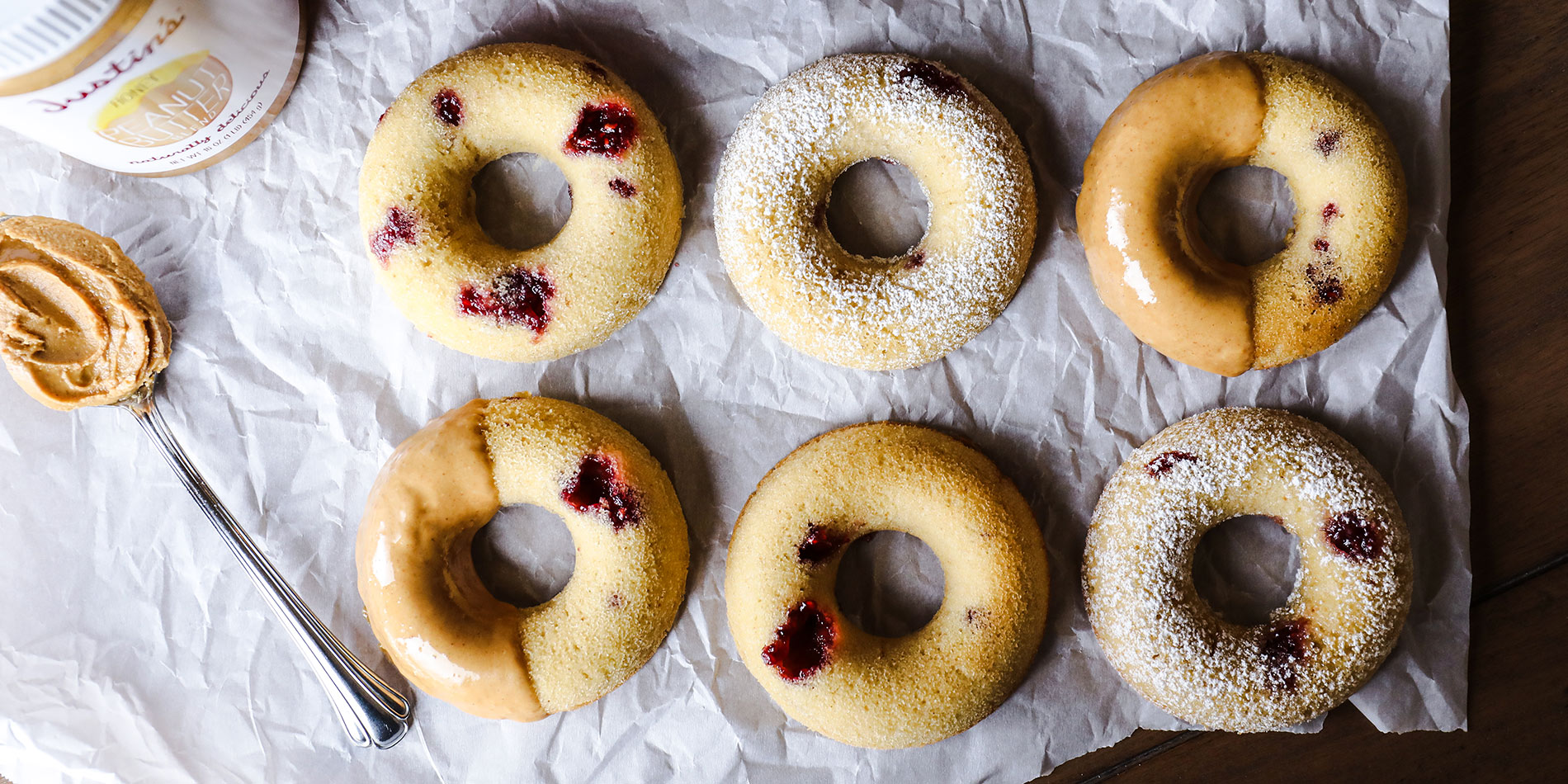 Gluten-Free PBJ Doughnuts on crinkled white parchment paper with a spoonful of peanut butter on a wood background