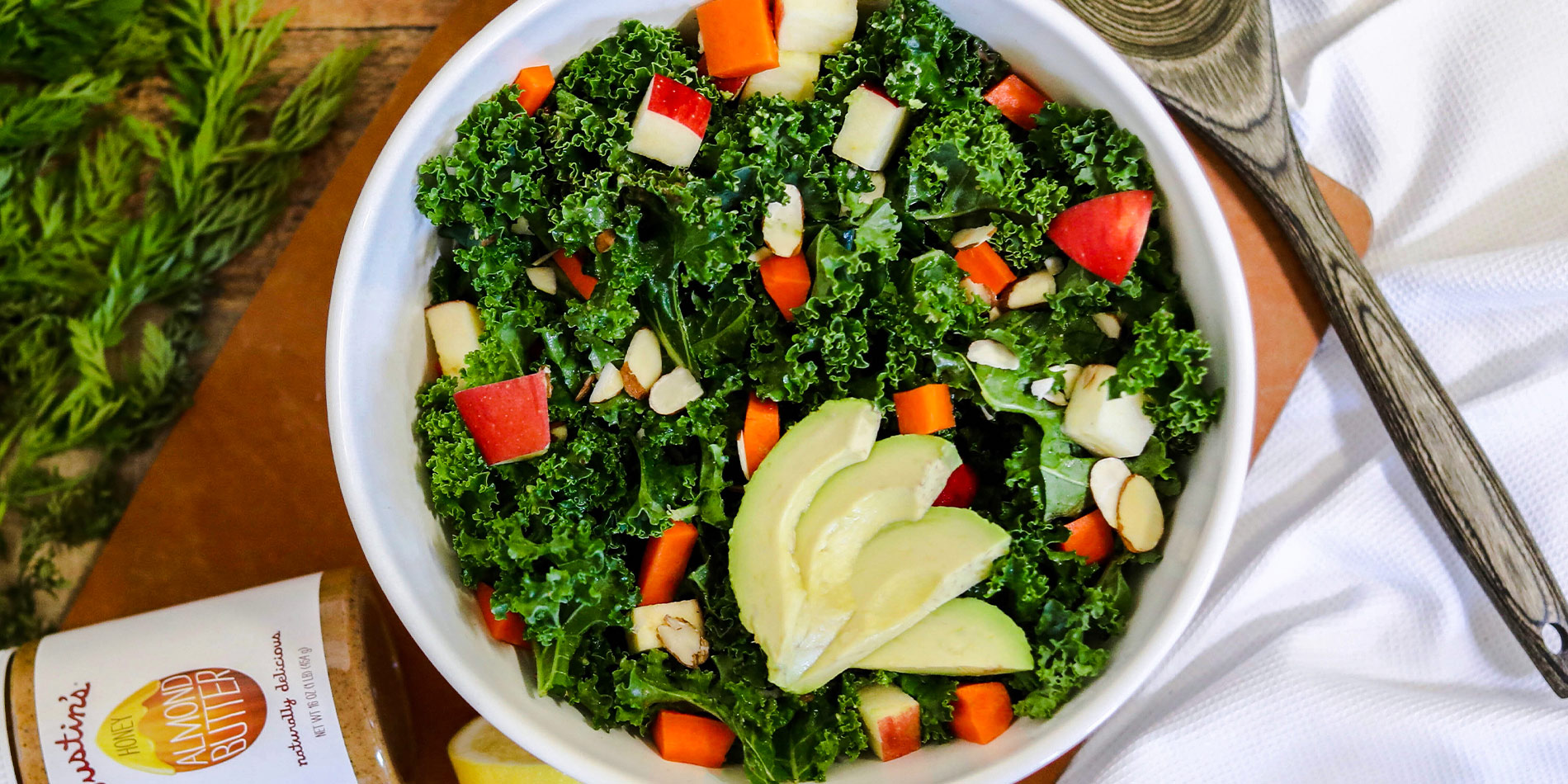 Massaged Kale Salad with Honey Almond Butter Vinaigrette in a white bowl with carrots and cut lemons in the wood background