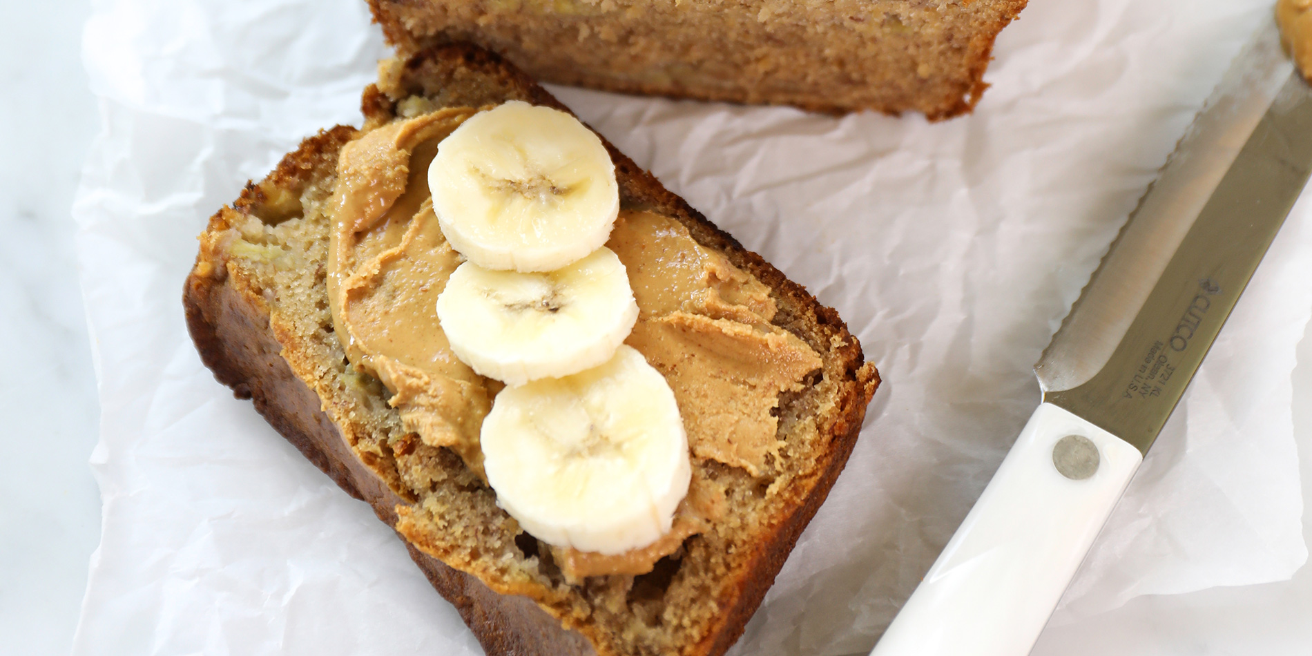 Peanut Butter Apple Banana Bread slices topped with banana slices and peanut butter on white parchment on white background