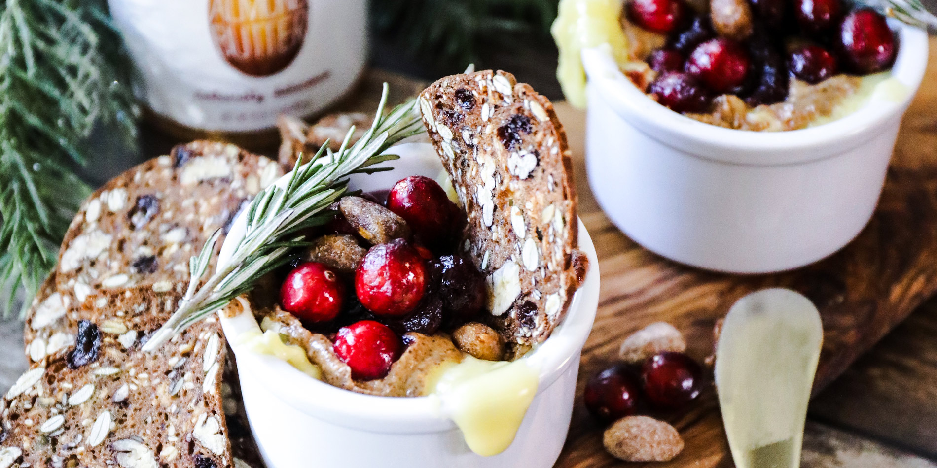 Cranberry Almond Butter Baked Brie in a small cup on a wooden serving tray with cranberries and pine on a wood background