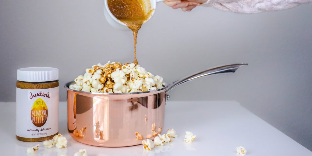 Honey Almond Buttered Popcorn in a pink pan with honey almond butter poured from top by a human hand on a white background