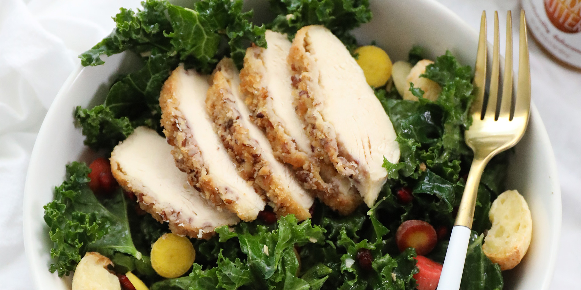 Kale Salad with Almond Butter Crusted Chicken on a white bowl with a golden fork on a white cloth on a white top