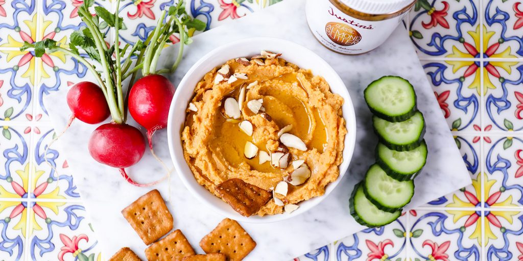 Sweet Potato Almond Hummus on a white bowl with cucumber slices, beets, crackers on a floral patterned tile background