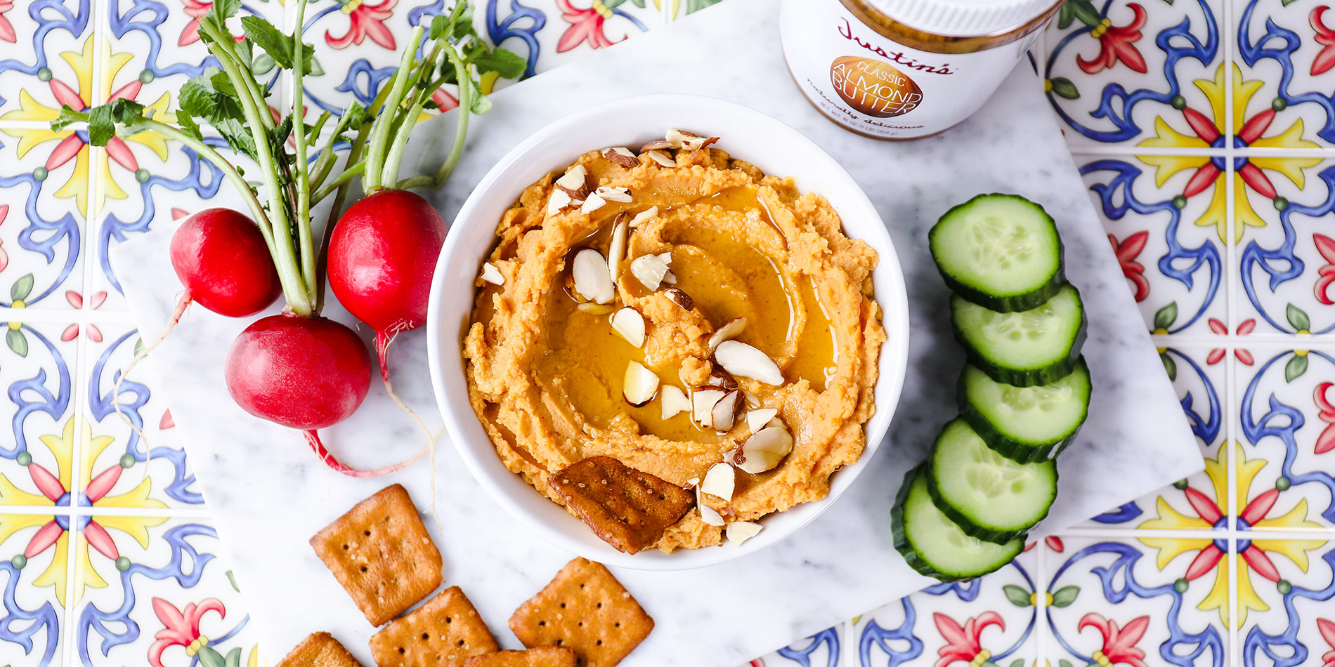 Sweet Potato Almond Hummus on a white bowl with cucumber slices, beets, crackers on a floral patterned tile background