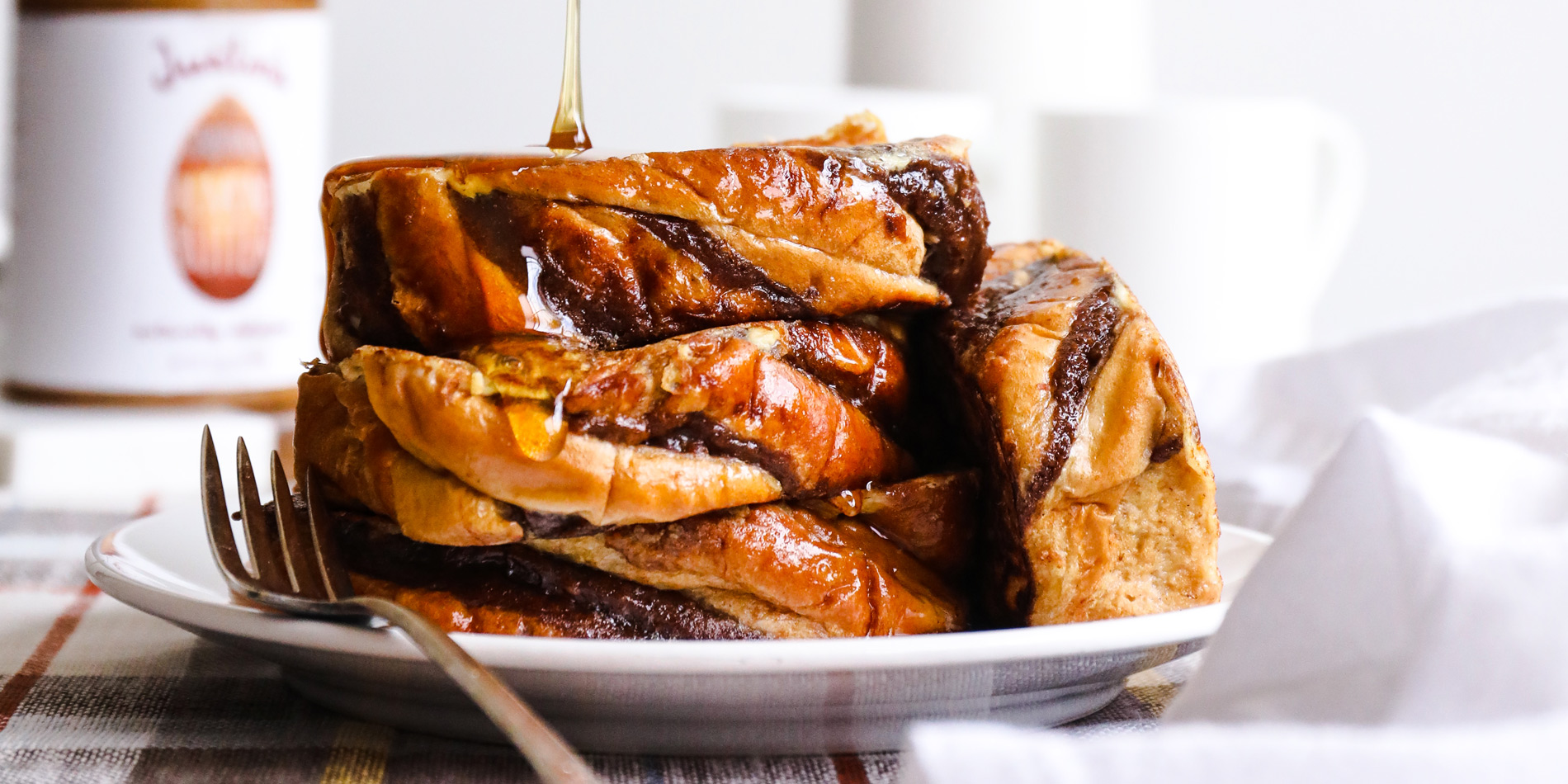 Almond Butter French Toast stacked on a round plate with a brass fork on the side, with a glaze poured from the top