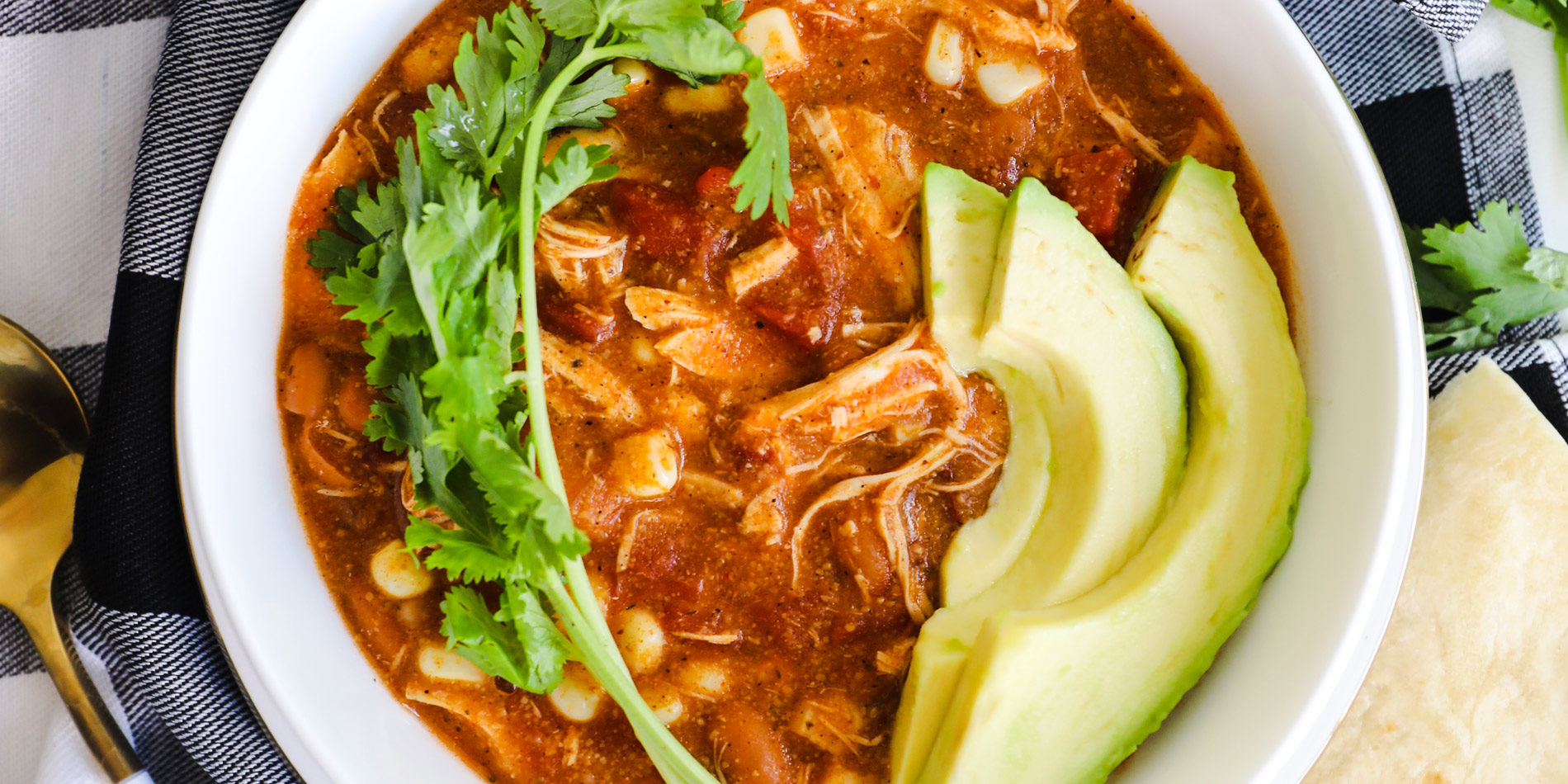 Chipotle Chicken Enchilada Soup in a white bowl with avocado and cilantro and tortillas on a checkered cloth