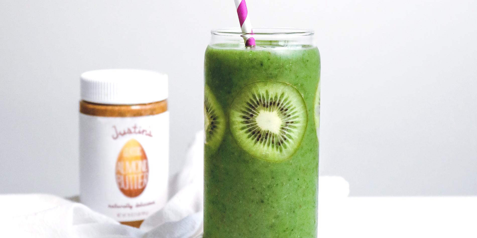 Justin’s Ultimate Green Drink in a clear cup with a striped straw with kiwis on a white background