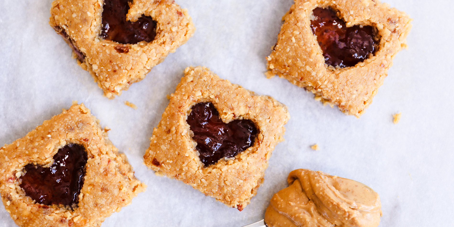 No Bake PB & J Bars with heart-shaped jelly center with a spoonful of peanut butter on white background with crumbs