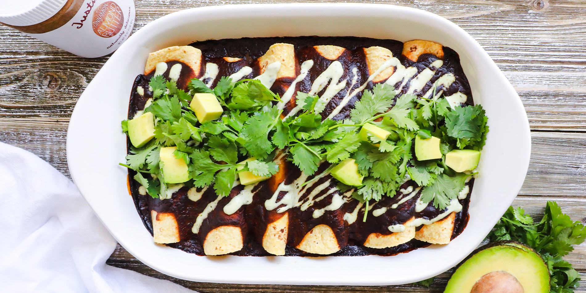 Vegetable Enchiladas with Cinnamon Almond Butter Mole in a white bowl with an avocado on top of cilantro on a wood background