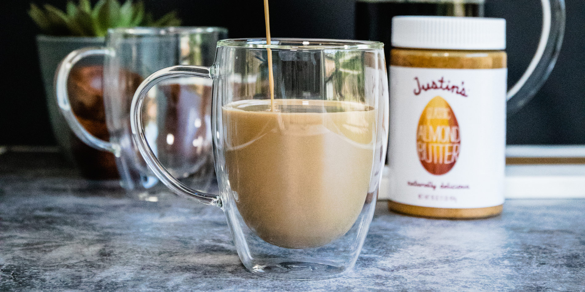 Almond Butter Bulletproof Coffee dripped down from a metal jug to a transparent cup on a grey marble tabletop and black wall