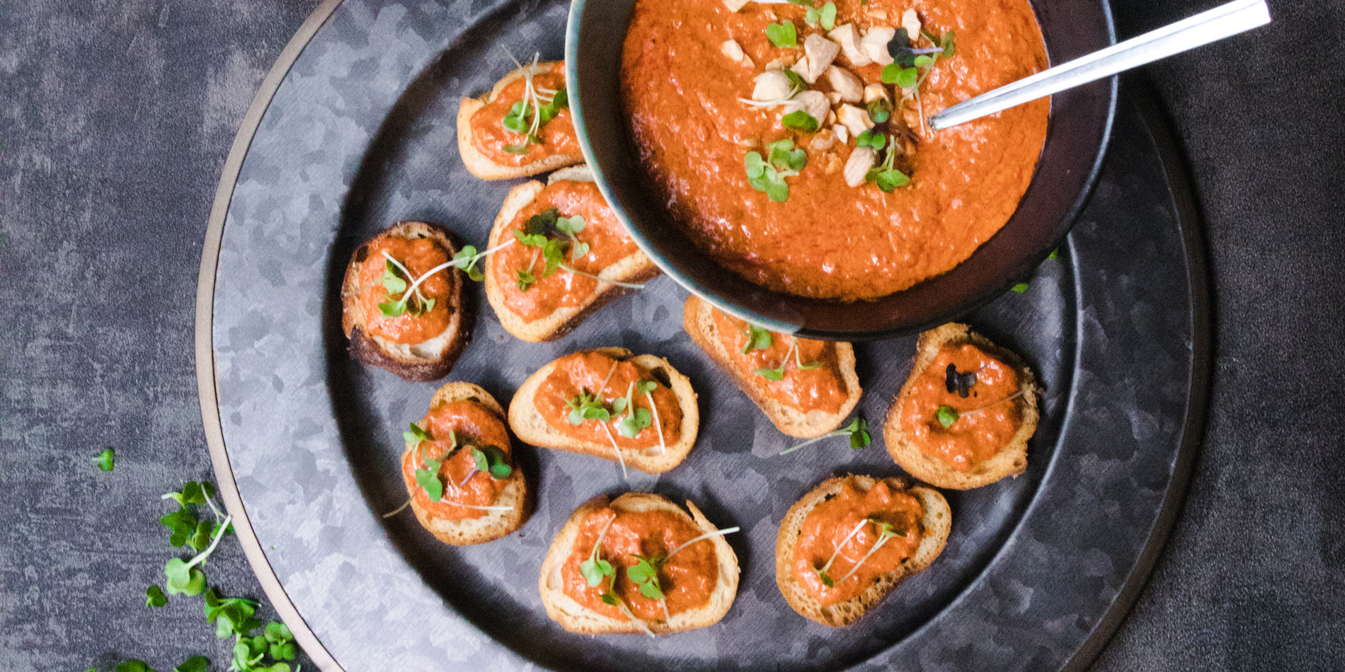 Almond Butter Romesco in a two-colored bowl on a gray plate filled with small bread slices with Romesco on them