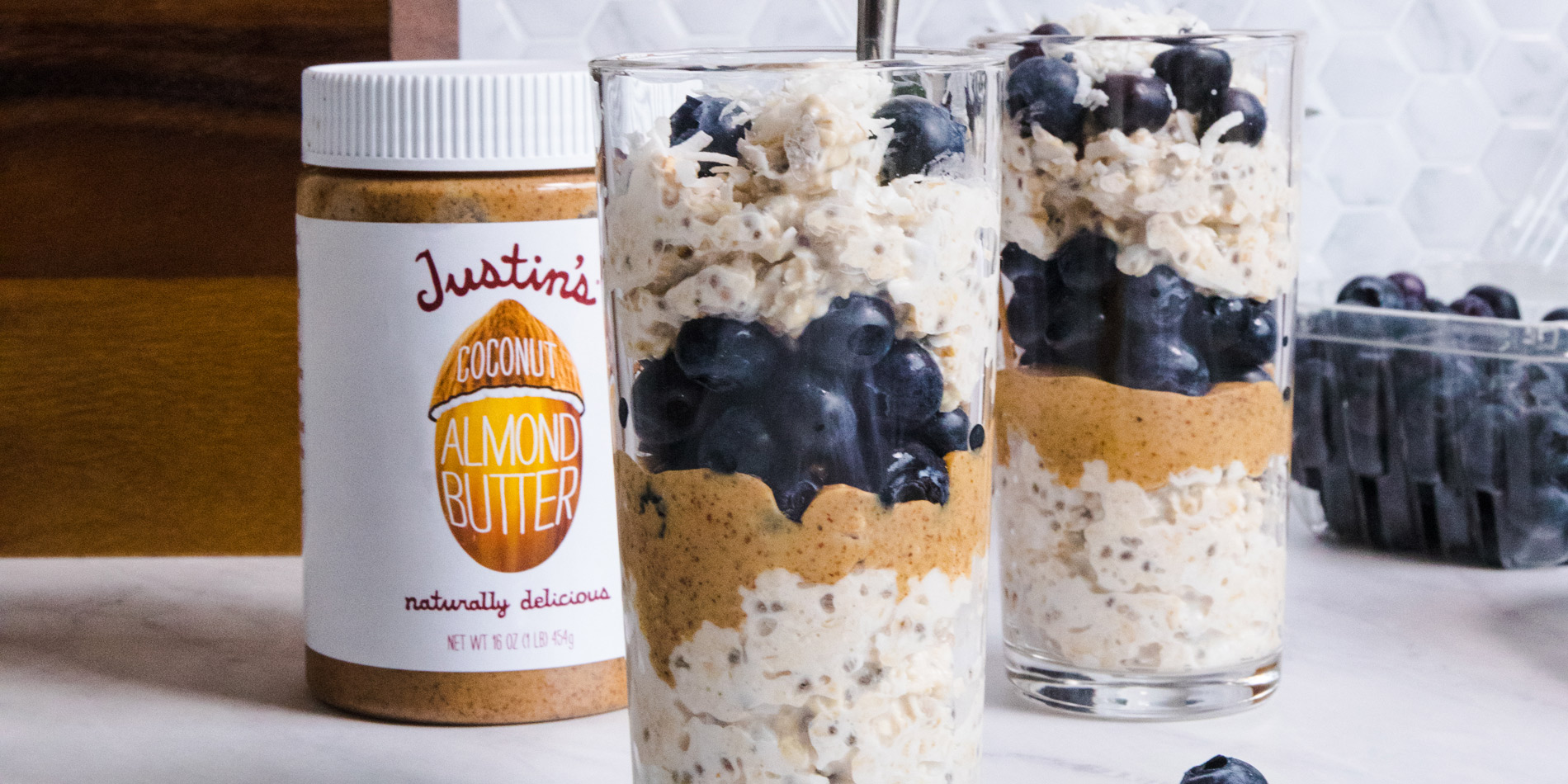 Coconut Almond Butter Overnight Oats in a clear cup with a bamboo straw, and wooden baking utensils in the white background