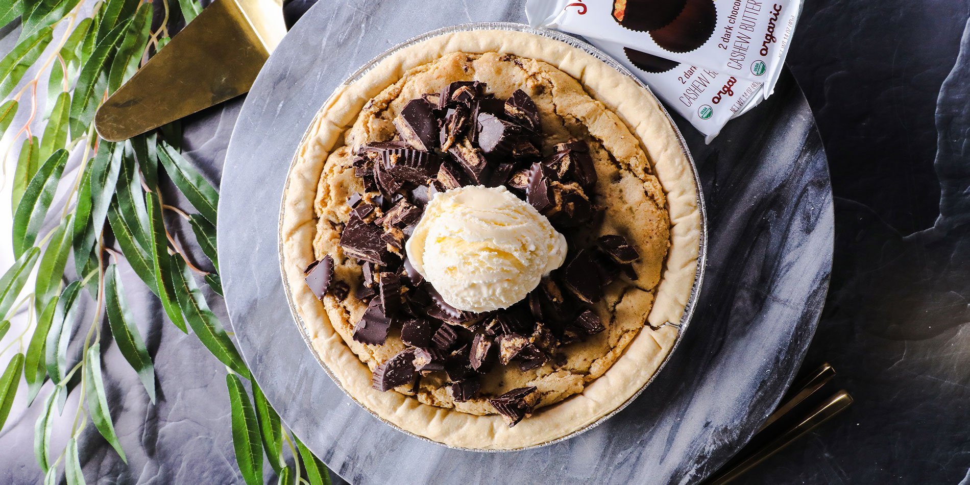 Nut Butter Cup Cookie Pie on a gray marbled tray on a gray background with leaves and golden pie server with wooden handle