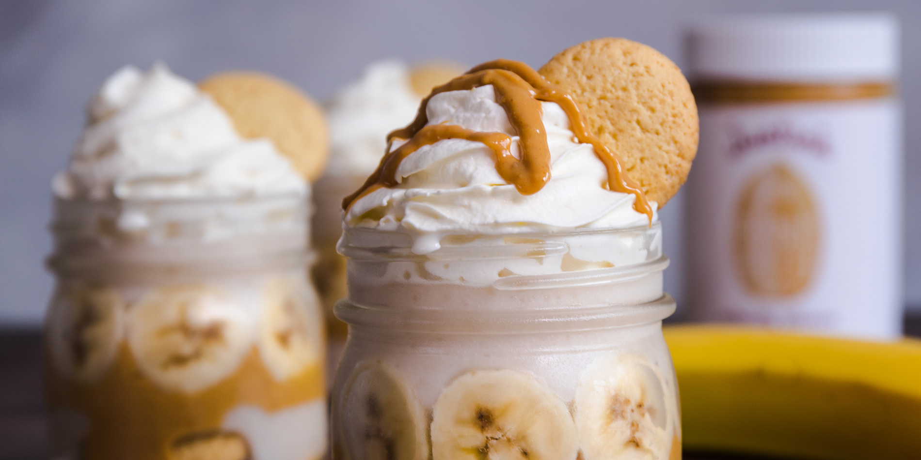 Peanut Butter Banana Pudding with whipped cream and peanut butter drizzle with cookies on a wooden top with white background