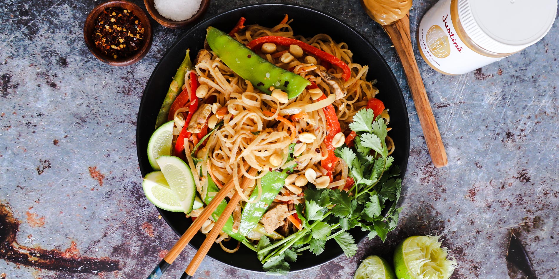 Thai Peanut Noodles in a black bowl with a spoonful of peanut butter on the side on a gritty gray background with lime slices