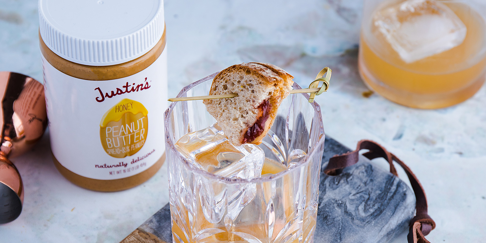 Honey Peanut Butter Old Fashioned with a piece of PB & J sandwich on a toothpick that is placed on the rim of the clear cup