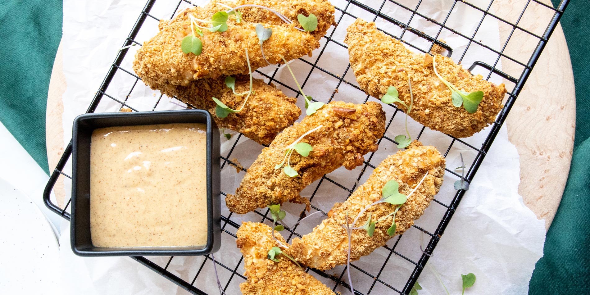 Pretzel Crusted Chicken Tenders with Maple Almond Dijon Dipping Sauce on a black cooling rack on top of a round wooden tray