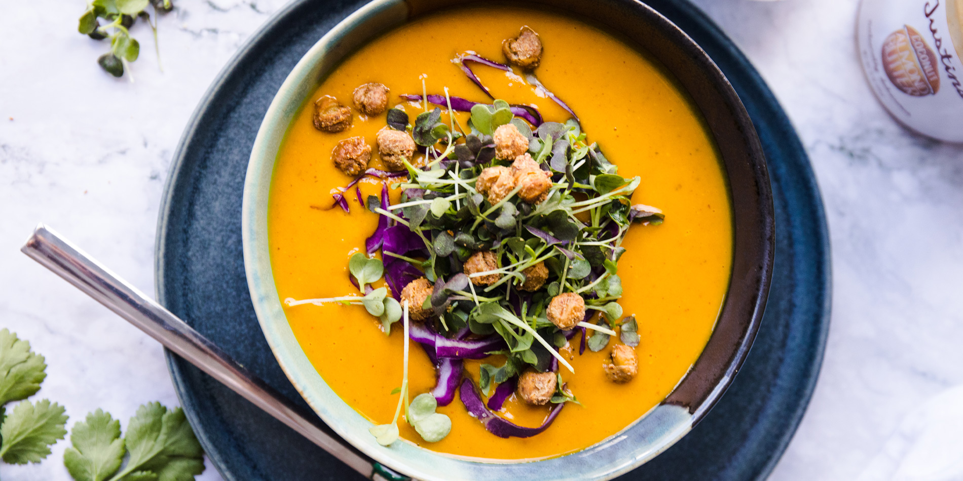 Thai Coconut Curry Soup in a two-colored bowl on a grey round plate on a white marble tabletop with cilantro leaves