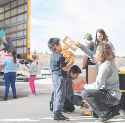 Justin crouching in front of a kid holding Justin's packaged goods as other people unload the truck