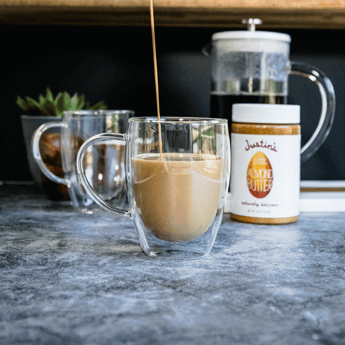 Almond Butter Bulletproof Coffee dripped down from a metal jug to a transparent cup on a grey marble tabletop and black wall