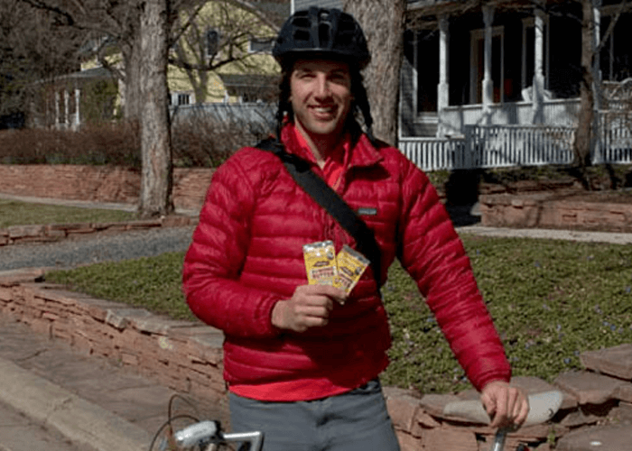 Outdoor shot of Justin wearing a bicycle helmet, holding products in hand while standing beside his bike